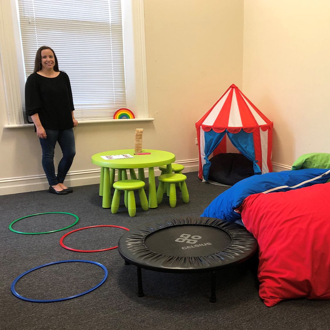 A kid's play room with a mini trampoline, a small text, some hoops, bean bags, and a table.
