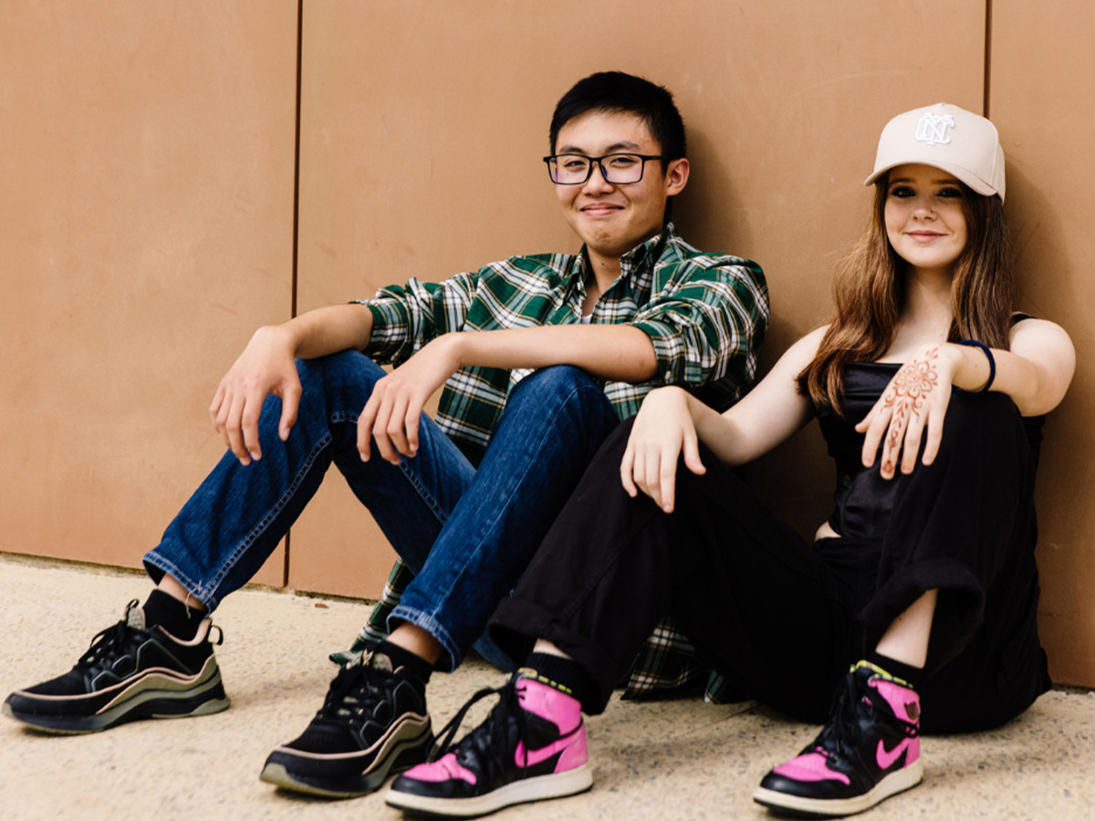 Two young people sitting on the ground against a wall, both smiling at the camera.